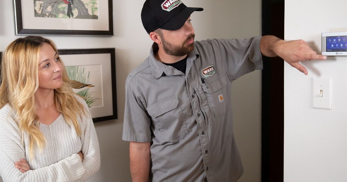 HVAC technician in a grey uniform and cap explaining thermostat settings to a female homeowner in a cozy room, demonstrating home temperature control system